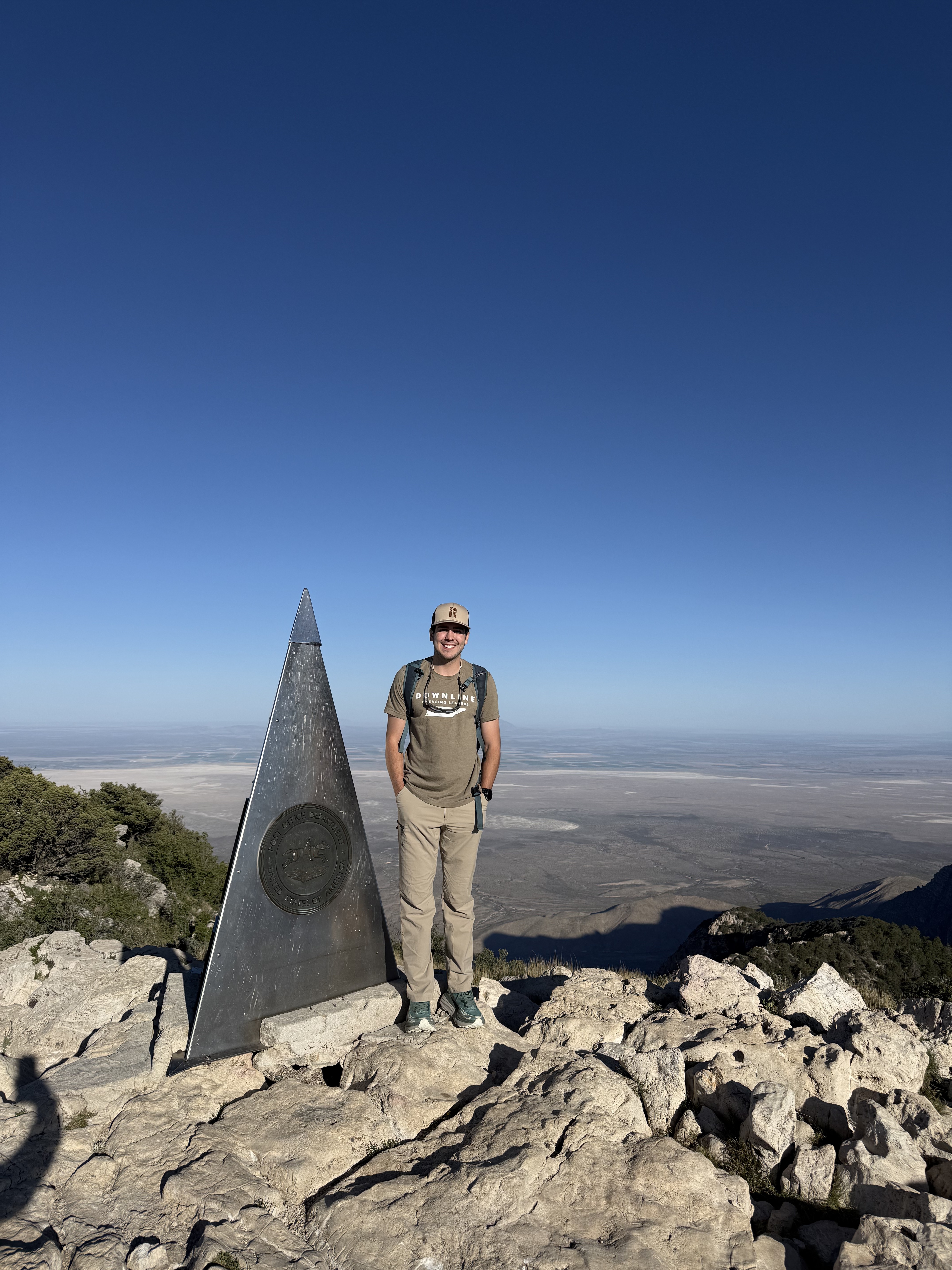 Christopher Ochoa standing beside a summit marker at the top of a mountain
